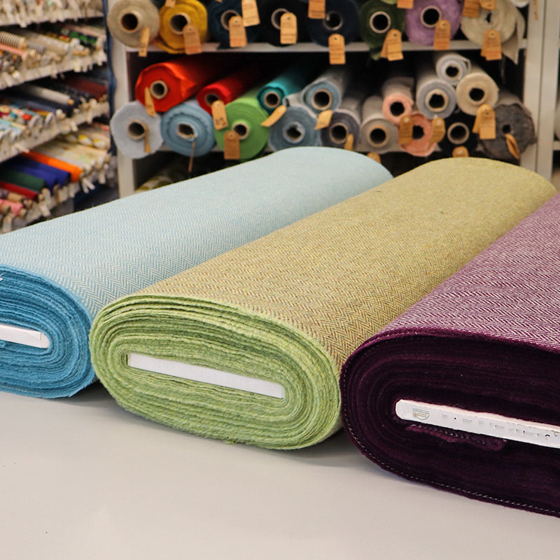 Three rolls of wool herringbone fabric in blue, green, and dark purple
