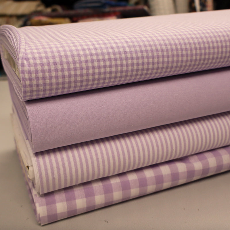 A stack of four lavender and white cotton fabrics with different patterns, including solid, stripes, and gingham